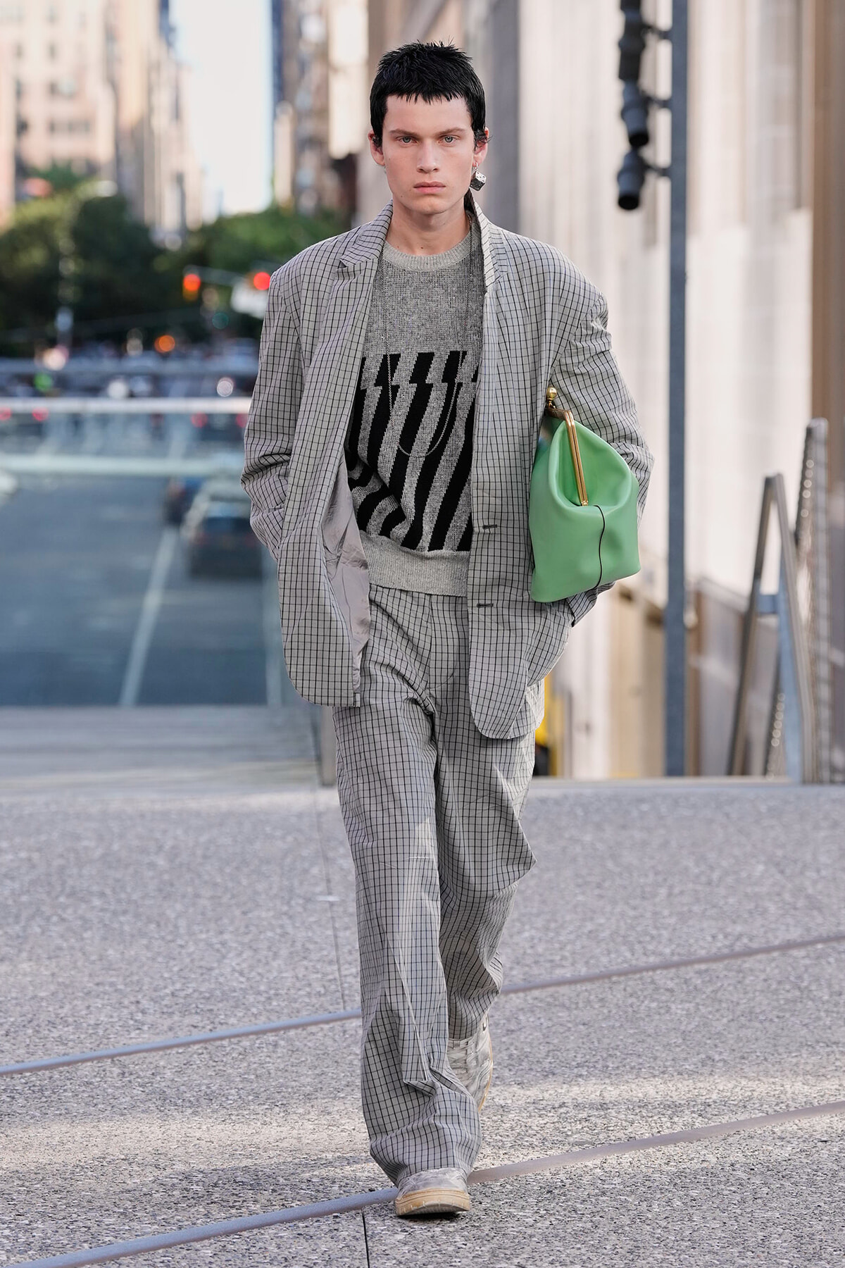 Fashion model in a gray checkered suit and patterned sweater walking down a city street, carrying a green handbag under his arm.