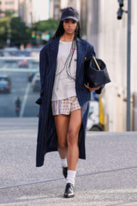 Person walking on city street wearing a navy pinstripe long coat, graphic t-shirt, and patterned shorts, with a cap and white socks, carrying a black bag.