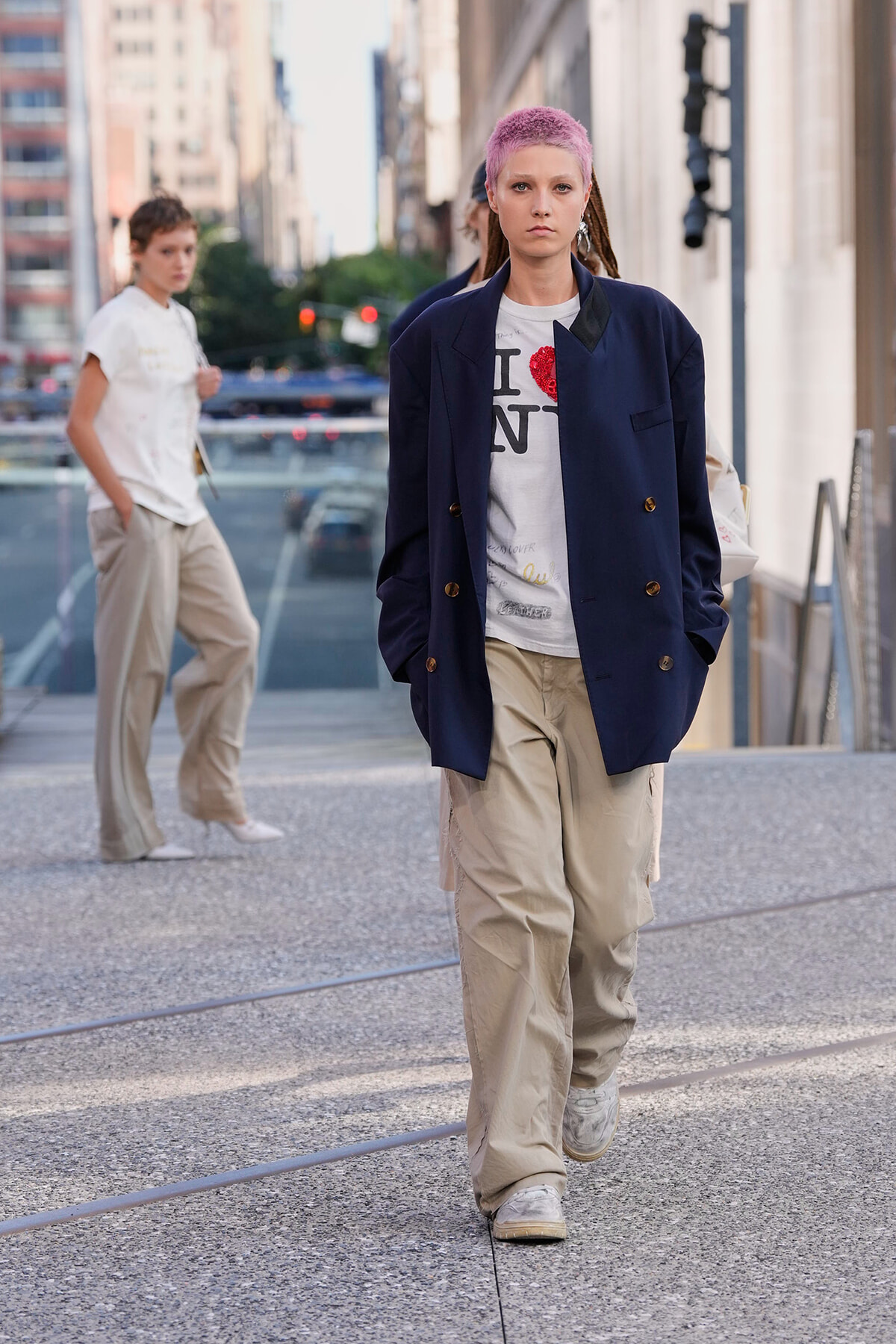 Person with pink hair wearing a navy blazer and beige cargo pants walks toward the camera on a city sidewalk.