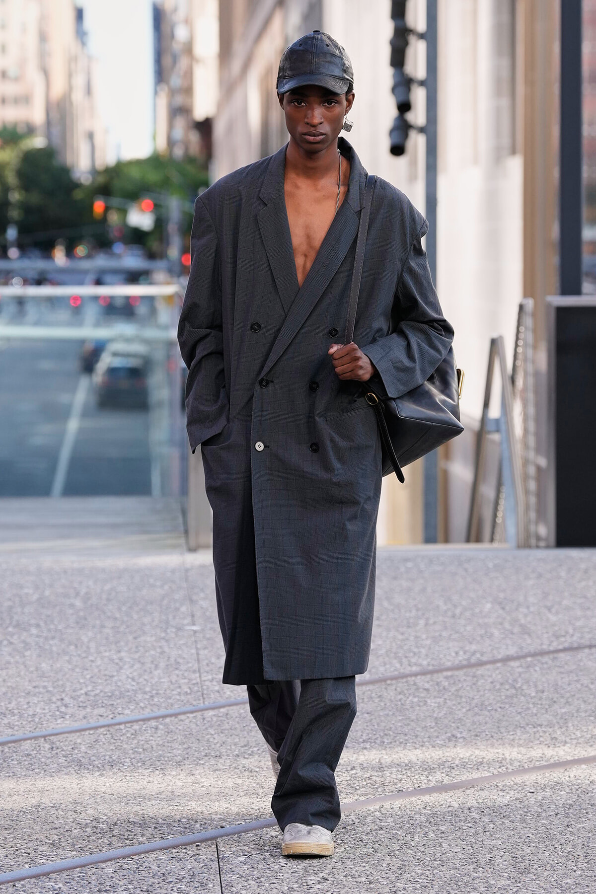Person walking on a city sidewalk in an oversized gray double-breasted suit, cap, and carrying a black shoulder bag.