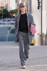 Woman in a gray plaid suit with a black zigzag sweater, cap, and pink bag walking down a city street.