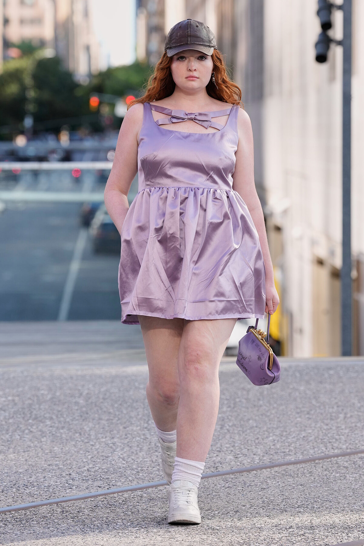 Red-haired woman walking on a city street in a lavender dress with a bow detail, wearing a cap and white sneakers, and carrying a small purple purse.