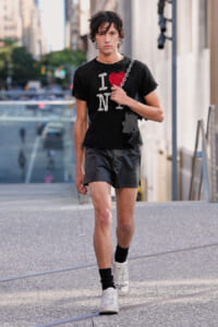 Man walking on a city sidewalk in a black distressed I heart NY T-shirt, black leather shorts, white sneakers, and a chain bag.",