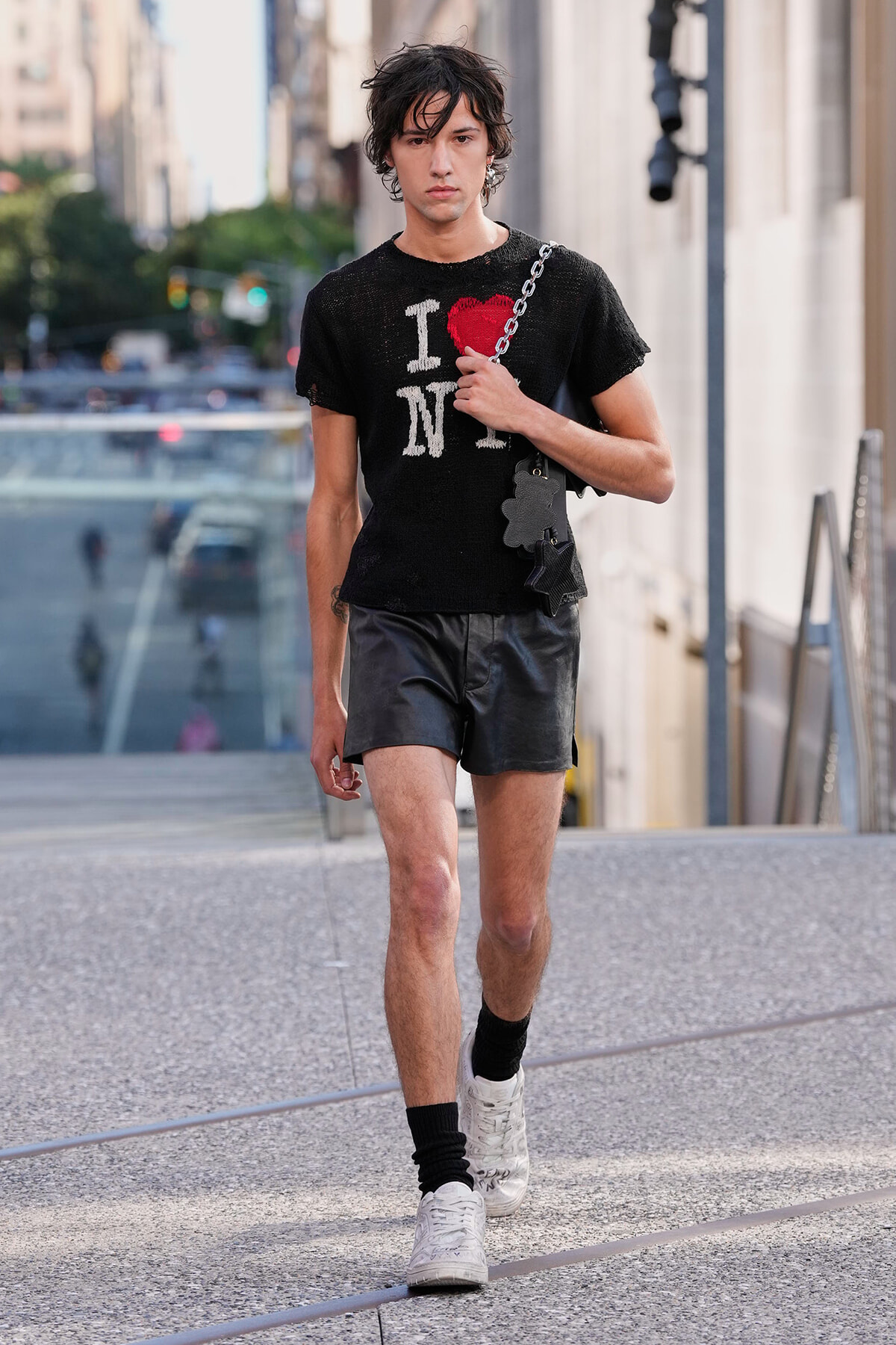 Man walking on a city sidewalk in a black distressed I heart NY T-shirt, black leather shorts, white sneakers, and a chain bag.",