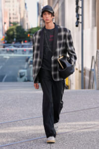Young man in a gray plaid blazer over a black tee and pants, cap, and white sneakers walking city streets with a black bag.