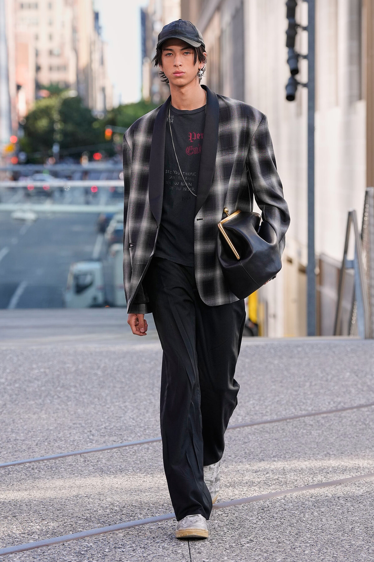 Young man in a gray plaid blazer over a black tee and pants, cap, and white sneakers walking city streets with a black bag.