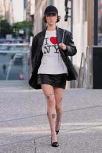 Person walking on a city sidewalk in a white I heart NY shirt, black leather jacket, black mini-skirt, cap, and glasses; tattoos on legs visible.