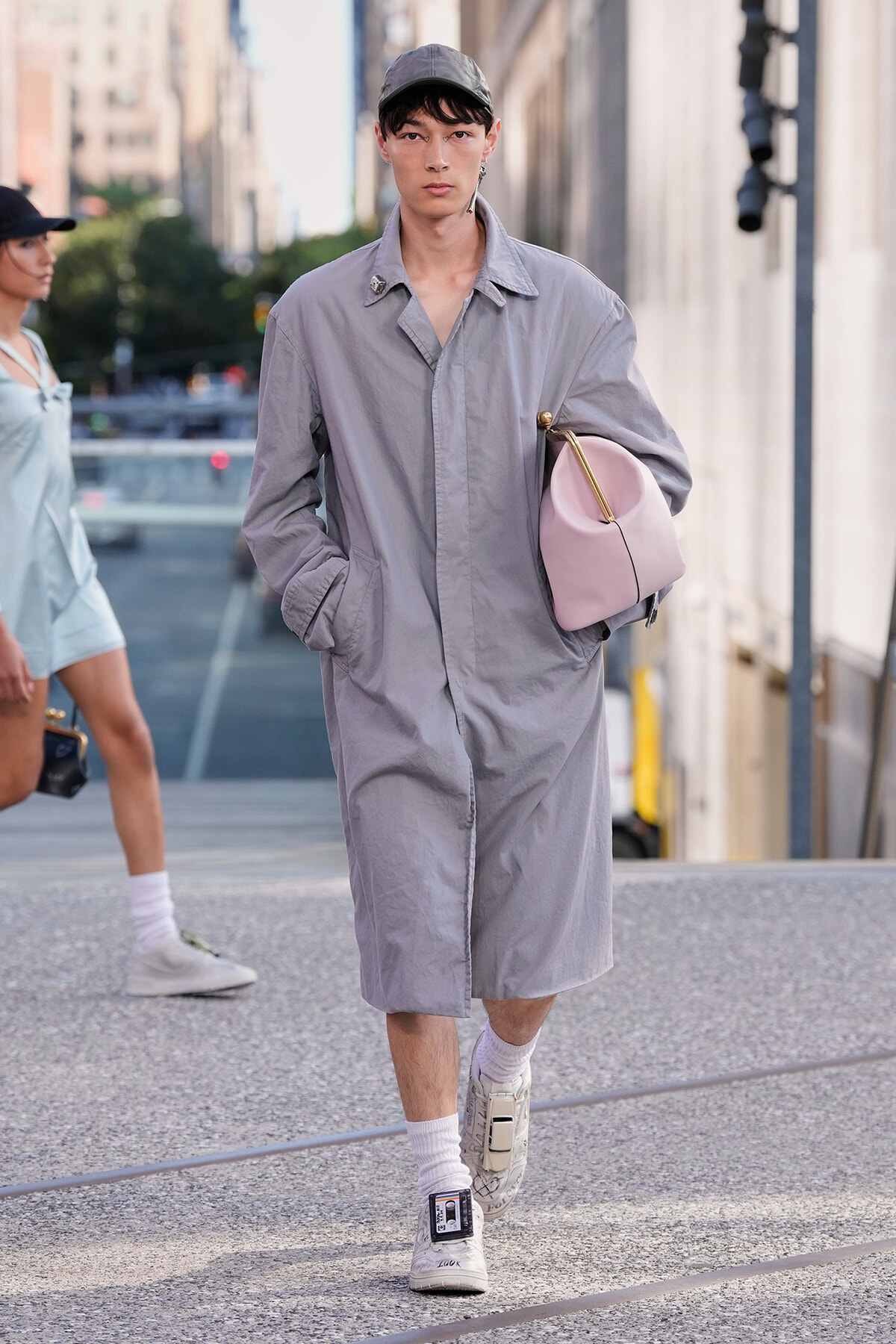 Model walking in a light gray oversized trench coat, cap, white sneakers, and a pink curved handbag in an urban street setting.