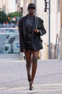 Young woman walks down a city sidewalk in an all-black outfit: blazer, cap, graphic tee, and black mini skirt, with a chain-strap bag over her shoulder.