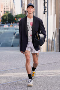 Person walking on a city street wearing a navy blazer over a graphic t-shirt, striped shorts, a cap, and carrying a black bag. Tattoos visible on legs.