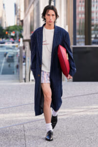Young man walking in city wearing a long navy pinstripe coat, white graphic T-shirt, striped shorts, white socks, and black loafers, carrying a red clutch under his arm.