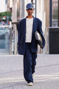 Person walking in a city street in a navy pinstripe suit, white T-shirt, blue cap, and carrying a black clutch.