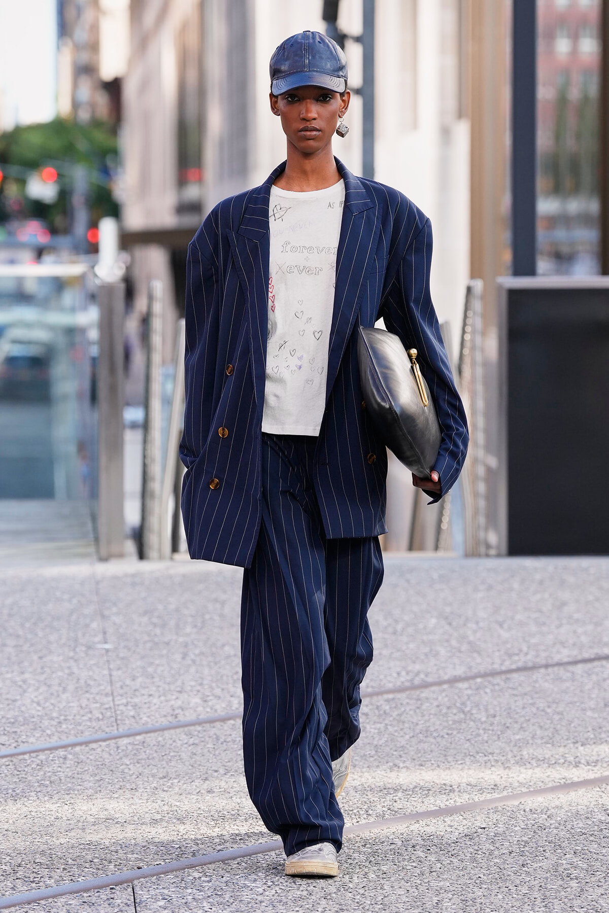 Person walking in a city street in a navy pinstripe suit, white T-shirt, blue cap, and carrying a black clutch.