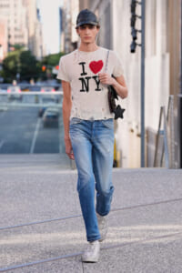 Model walking down a city street in an I ♥ NY sweater, faded jeans, white sneakers, and a dark cap, carrying a bag with a star charm.