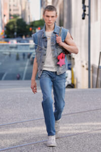 Young fashion model walking down a city street in a sleeveless denim vest, white tee, and blue jeans, carrying a brown bag with a pink star charm on the strap.