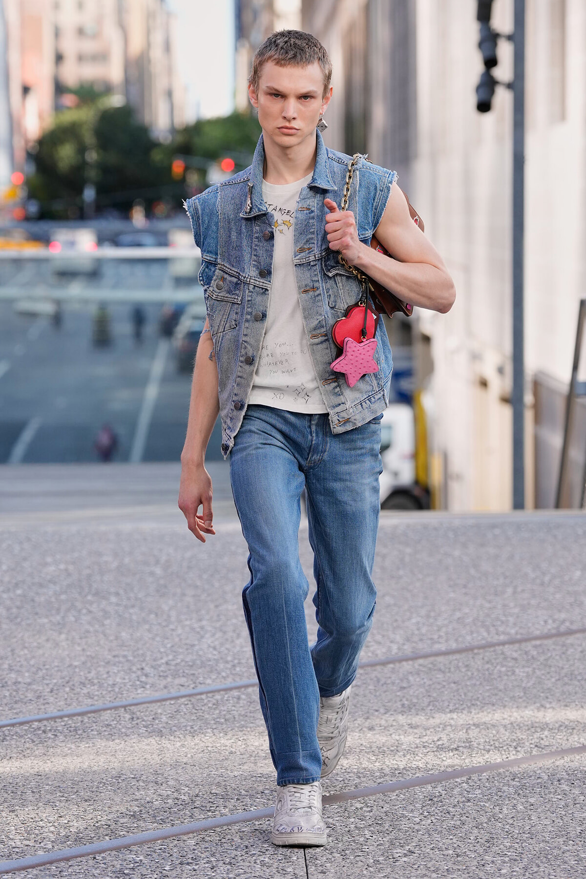 Young fashion model walking down a city street in a sleeveless denim vest, white tee, and blue jeans, carrying a brown bag with a pink star charm on the strap.