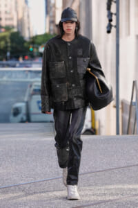 Person walking on a city sidewalk wearing a dark, patched black jacket, black leather pants, a cap, and carrying a large black bag over the shoulder or under arm.