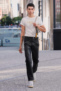 Male fashion model walking on a city street wearing a white graphic T-shirt, black leather pants, and white sneakers, carrying a chain-strap bag and accessories over the shoulder.