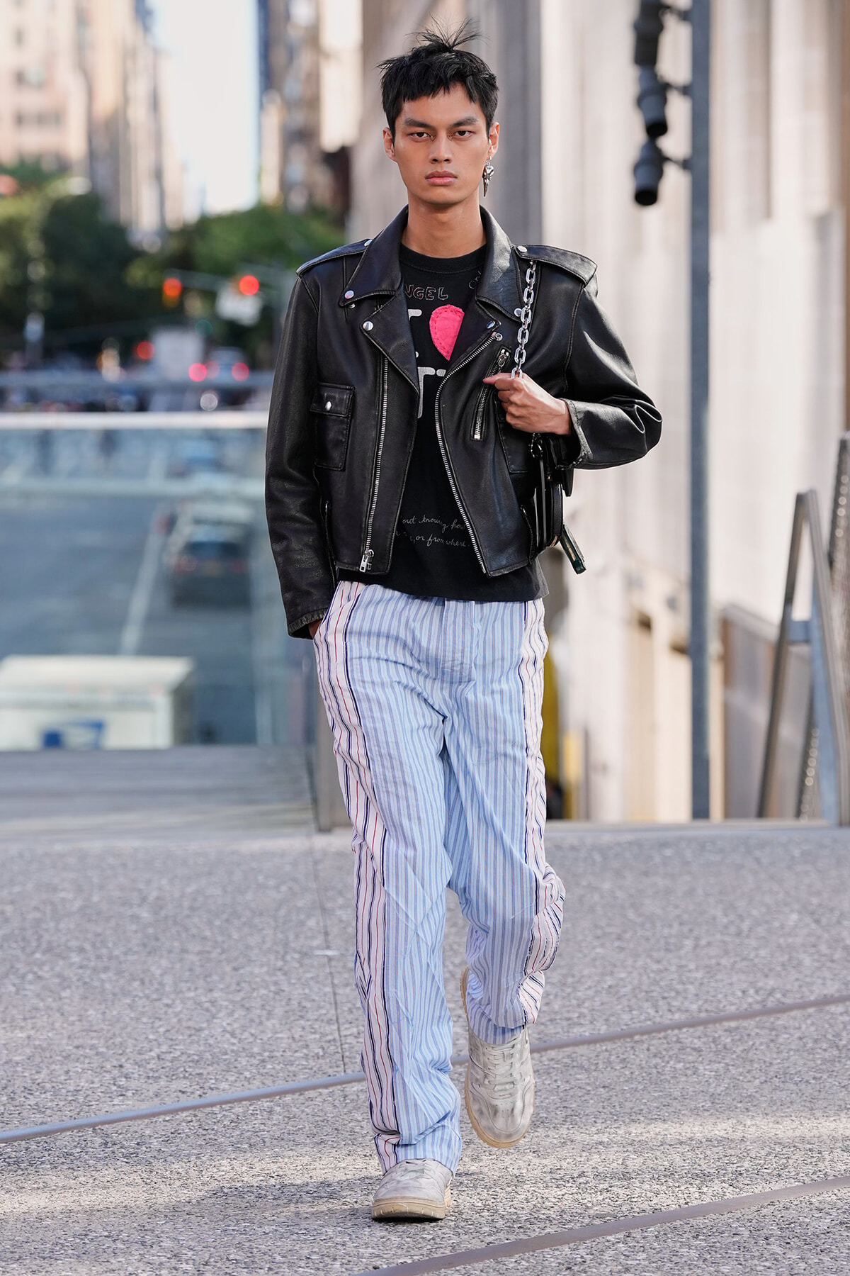 Young man walking on a city sidewalk in a black leather biker jacket, light blue striped pants, and white sneakers.