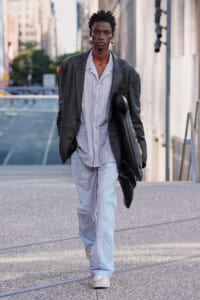 Man walking in an urban street in an oversized dark blazer over light striped loungewear, carrying a black bag.