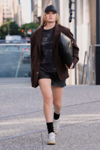 Street-style photo of a woman walking in an urban area, wearing a brown pinstripe blazer, black graphic tee, leather shorts, a black cap, and white sneakers while carrying a large black clutch.
