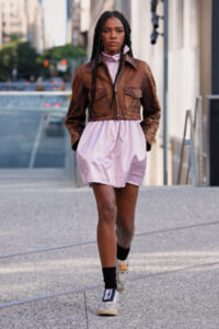 Woman walking on city street in a brown cropped leather jacket over a pale lavender dress; braided hair, sneakers, and black socks visible.
