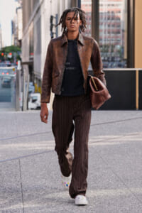 Urban street-style man walking on a city sidewalk in a brown leather jacket, black tee, pinstripe pants, white sneakers, and carrying a brown bag.