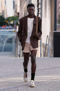 Young man walking on a city street in a brown pinstripe blazer, white graphic tee, and patterned shorts, with a tan crossbody bag on his shoulder.