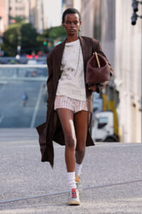 Model walking on a city street in a long brown pinstripe coat over a light graphic tee and striped shorts, carrying a burgundy handbag.