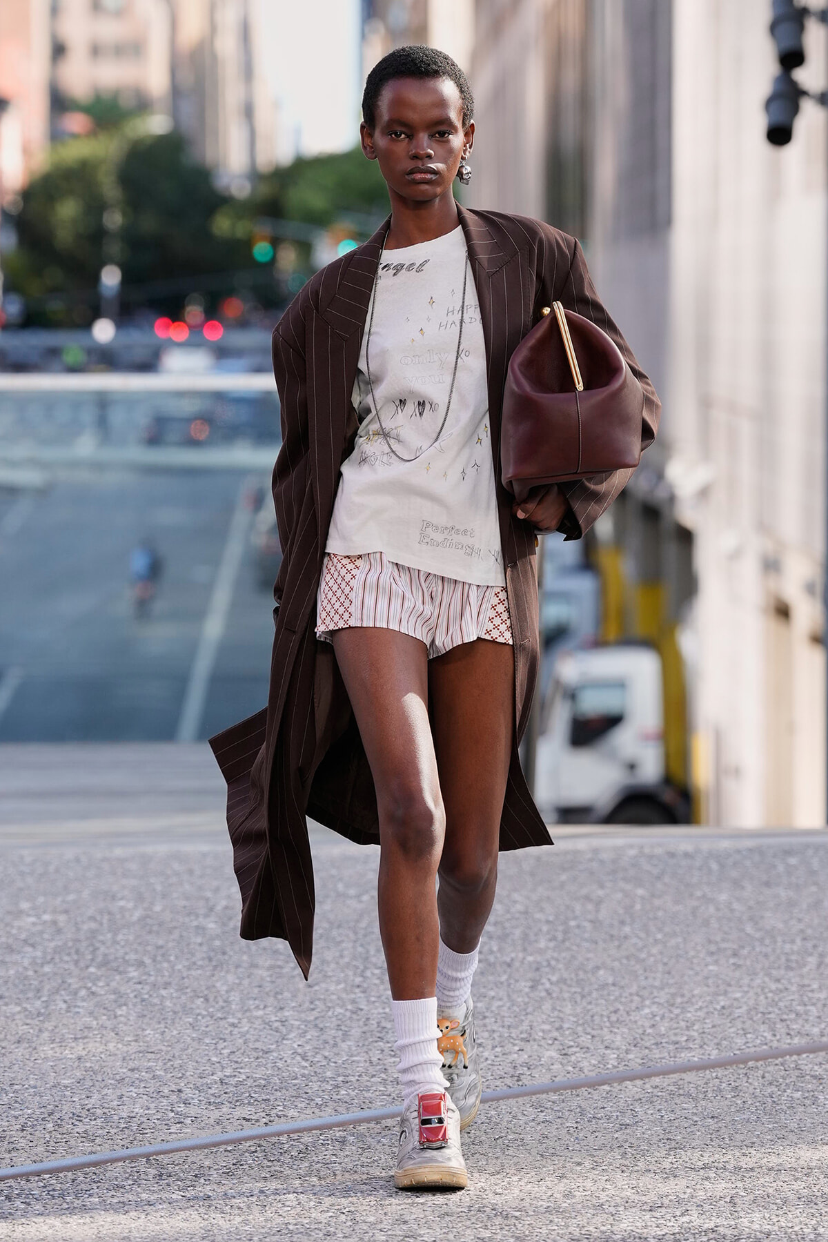 Model walking on a city street in a long brown pinstripe coat over a light graphic tee and striped shorts, carrying a burgundy handbag.
