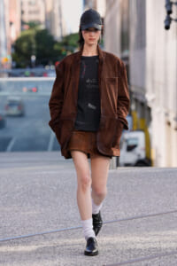Woman wearing an oversized brown velvet blazer and matching mini skirt walks on a city street, hands in pockets, wearing a black cap and oxford shoes.