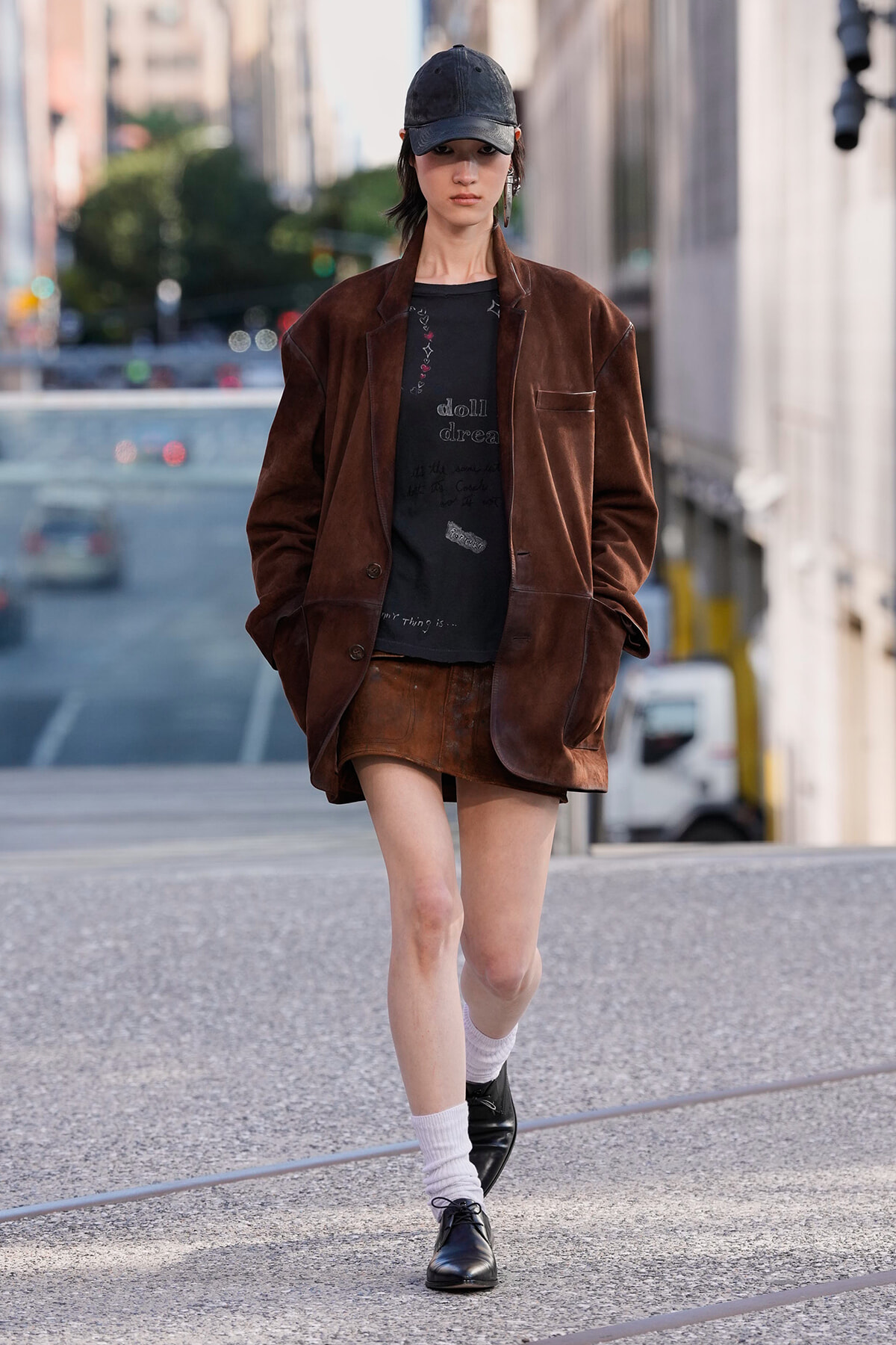 Woman wearing an oversized brown velvet blazer and matching mini skirt walks on a city street, hands in pockets, wearing a black cap and oxford shoes.