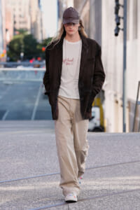 Young woman walking on a city street in a dark blazer, white graphic tee, and beige wide-leg pants with a cap.