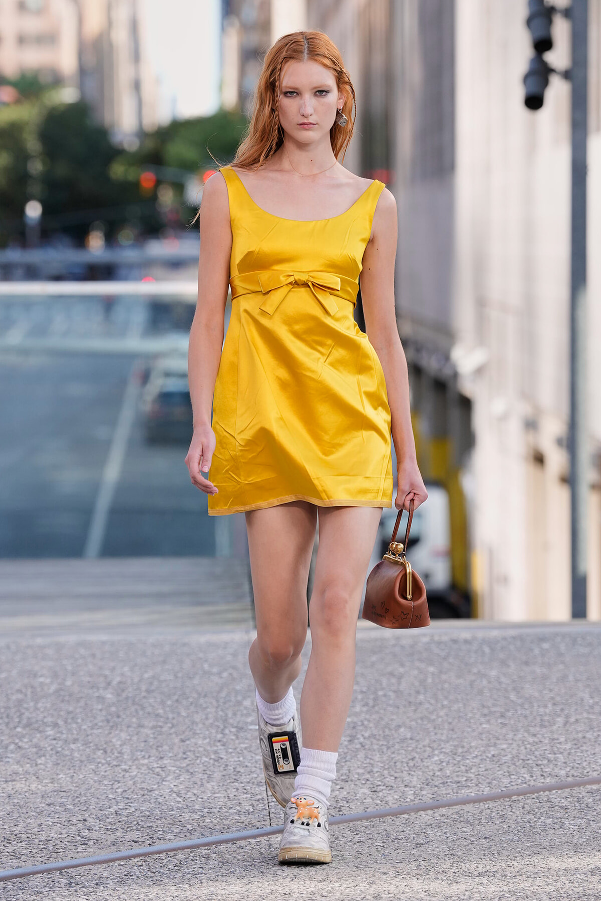 Model in a bright yellow satin mini dress with a bow at the waist walking on a city sidewalk, carrying a small brown purse.
