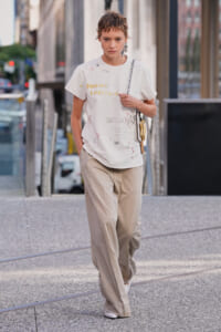 Model walking on a city sidewalk in a white graphic T-shirt and beige wide-leg pants, carrying a chain-strap bag over the shoulder.