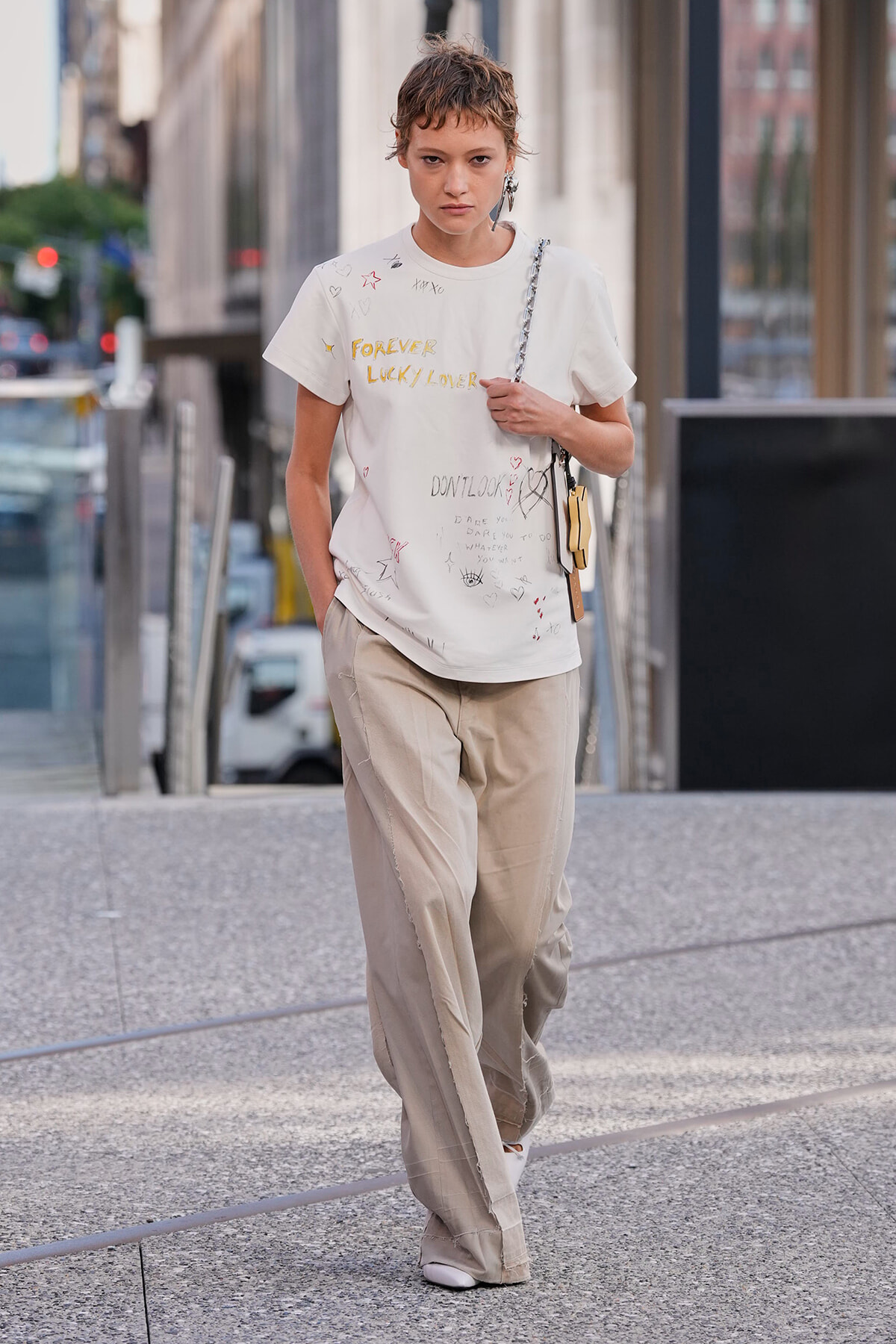 Model walking on a city sidewalk in a white graphic T-shirt and beige wide-leg pants, carrying a chain-strap bag over the shoulder.