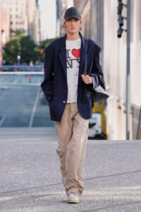 Person walking on a city street wearing a navy blazer, white I ❤️ NY tee, beige cargo pants, and a black cap with a shoulder bag over the shoulder