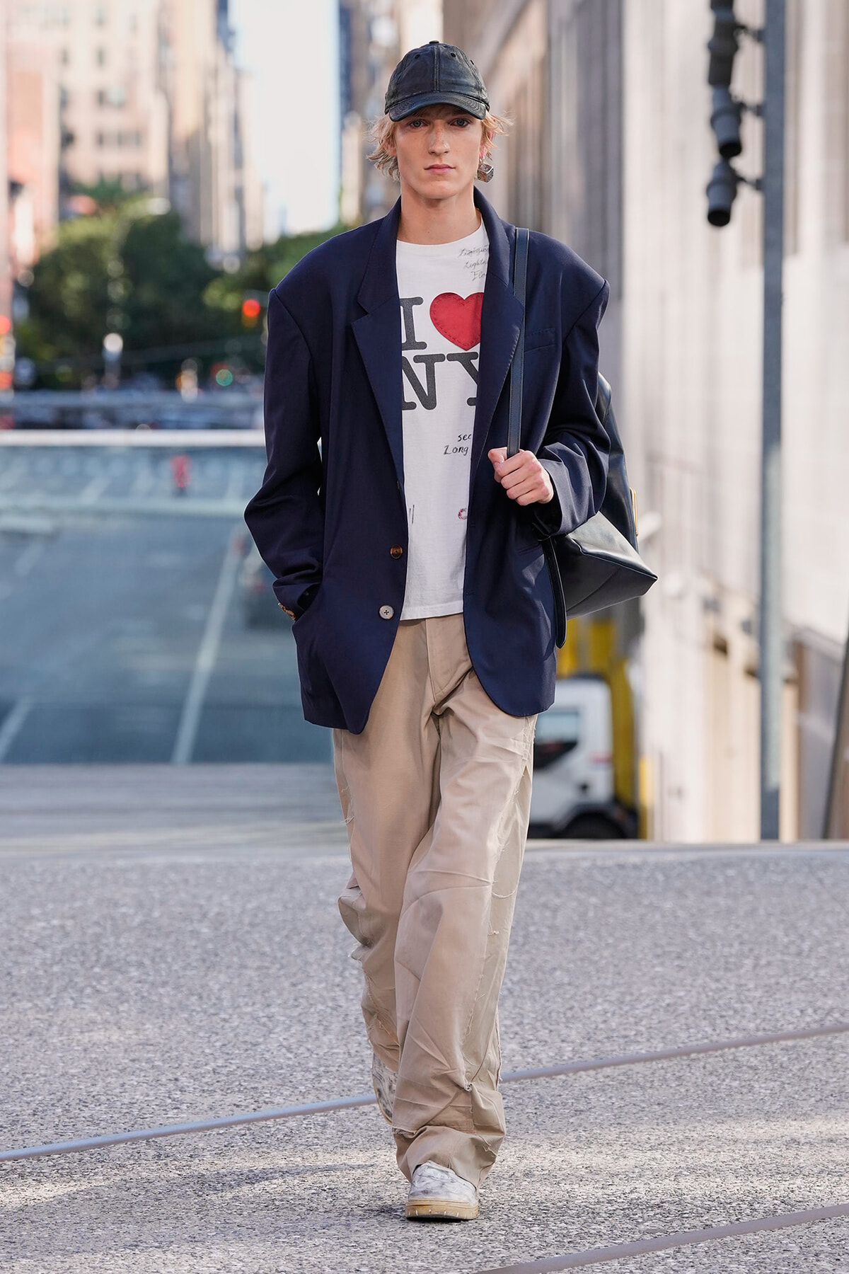 Person walking on a city street wearing a navy blazer, white I ❤️ NY tee, beige cargo pants, and a black cap with a shoulder bag over the shoulder