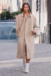 Person walking on a city sidewalk wearing a long beige trench coat, white sneakers, braided hair, and a white bag over the shoulder.