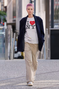 Person with short pink hair wearing an oversized dark blazer, white I ♥ NY shirt, and beige baggy pants walking on a city sidewalk.