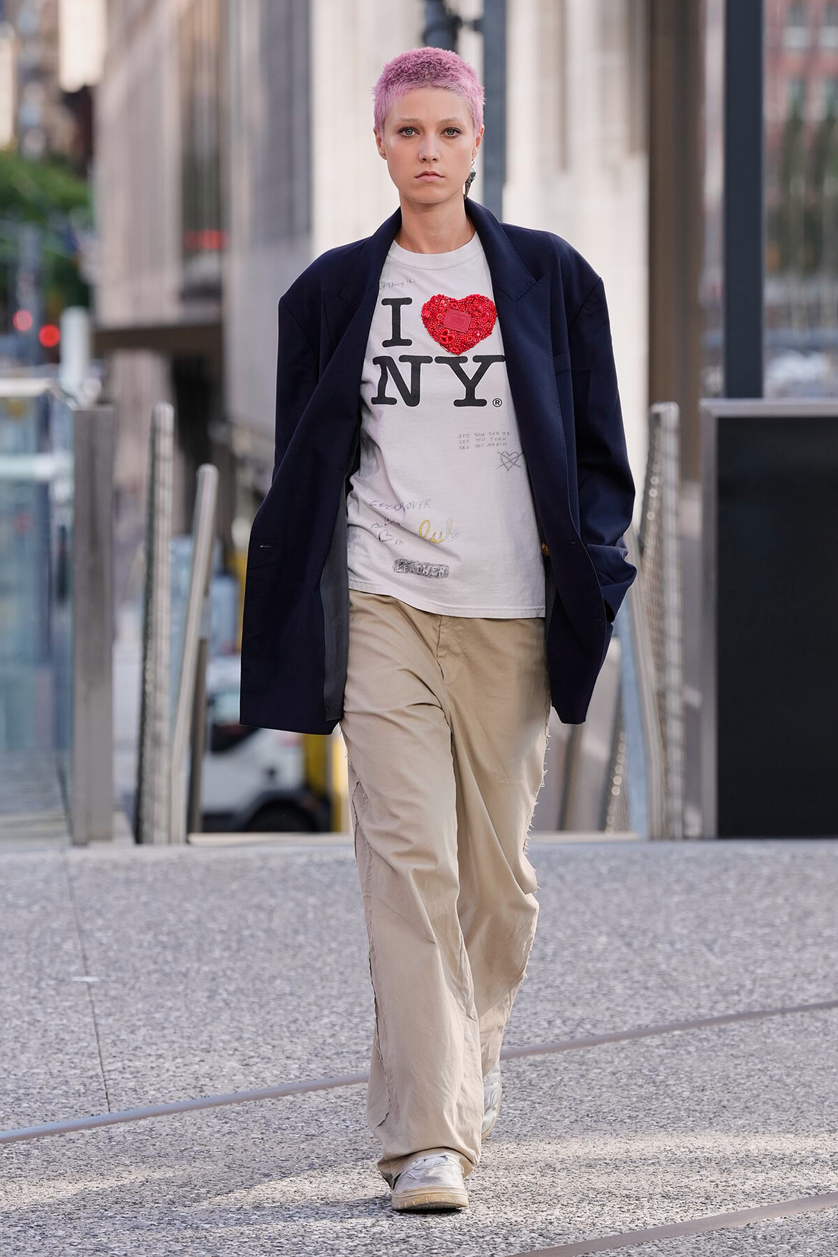 Person with short pink hair wearing an oversized dark blazer, white I ♥ NY shirt, and beige baggy pants walking on a city sidewalk.