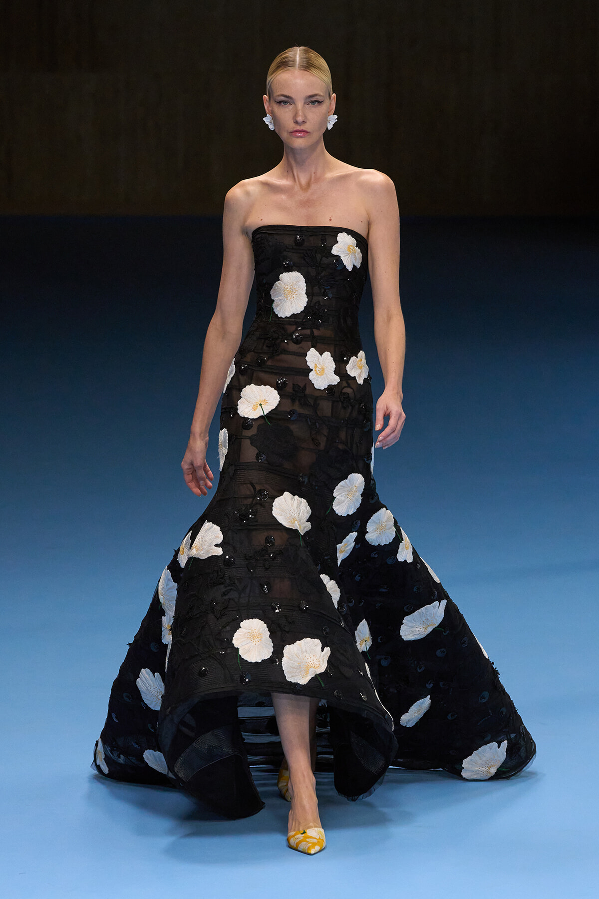 Model walking a strapless black gown with large white floral appliqués on a blue runway backdrop.