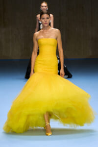 Model walks the runway in a strapless yellow mermaid gown with a voluminous tulle skirt and pearl embellishments at a fashion show.
