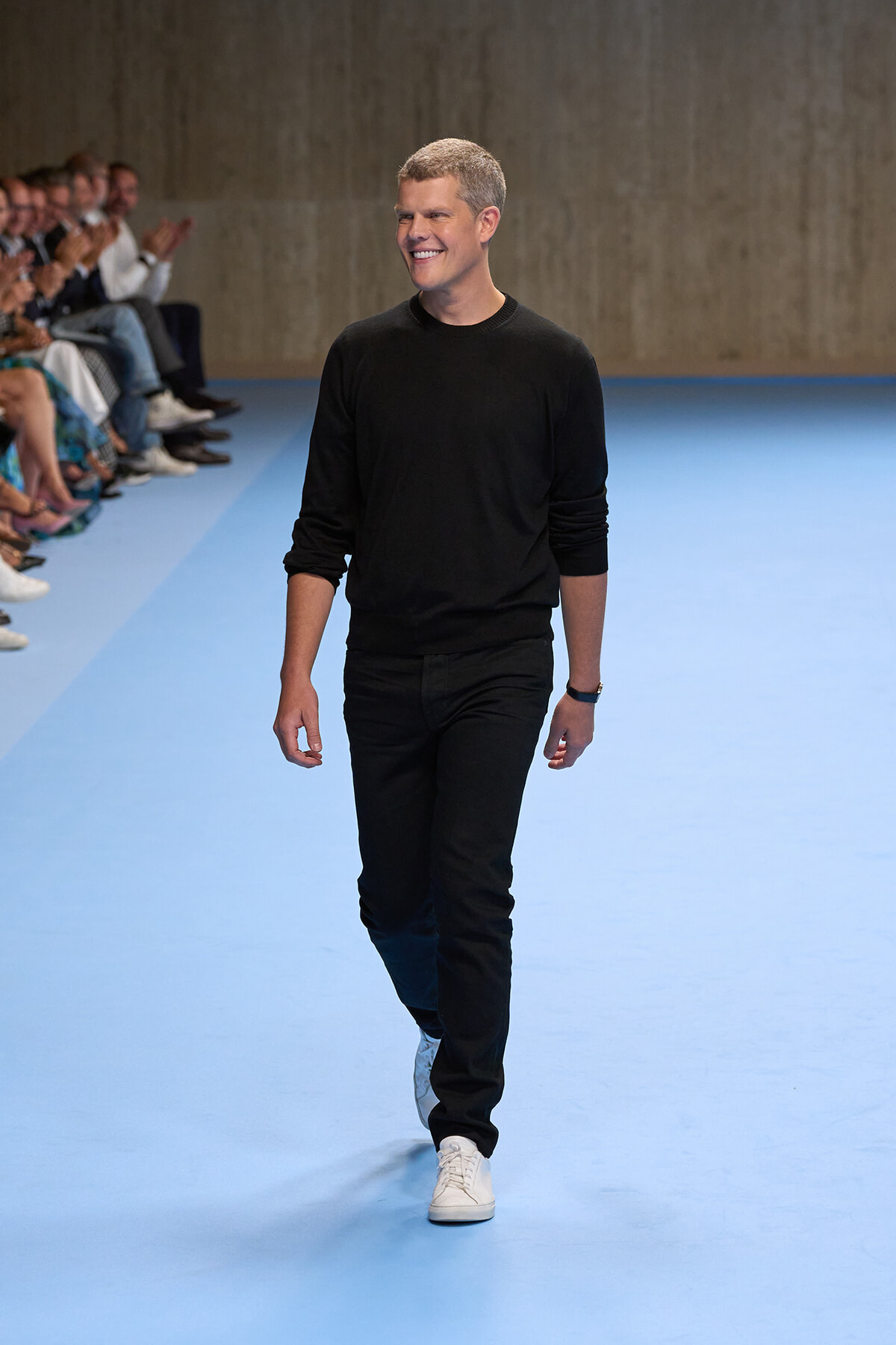 Male model walking down a blue runway in an all-black outfit (black sweater and pants) with white sneakers, audience on left applauding.