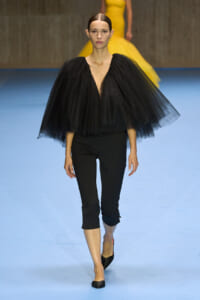 Model walks the runway in a dramatic black tulle cape over a deep V-neck top and fitted black cropped pants.