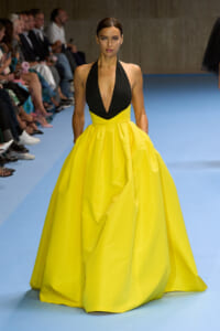 Model walks the runway in a black plunging halter top and a bright yellow full-skirt ball gown, with seated audience along the catwalk.