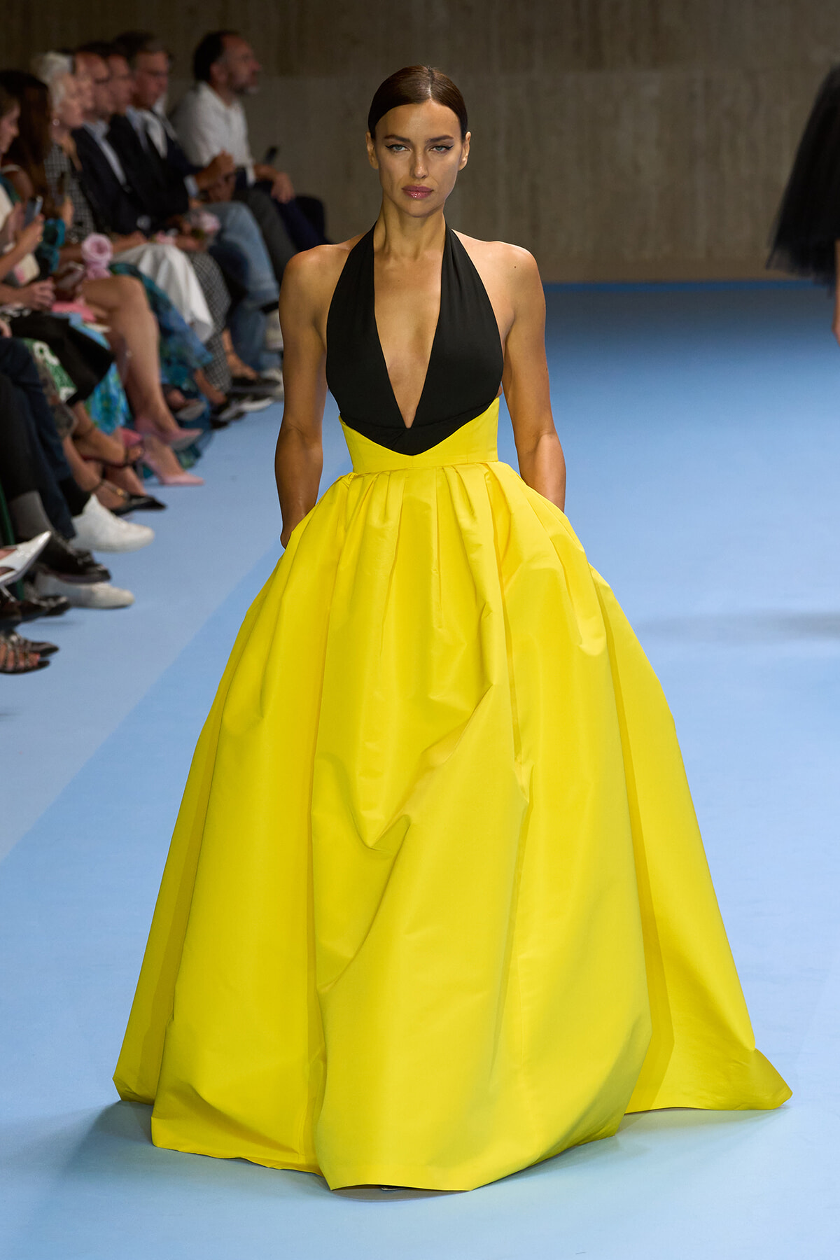 Model walks the runway in a black plunging halter top and a bright yellow full-skirt ball gown, with seated audience along the catwalk.