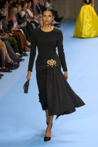 Model walks a fashion show in a black long-sleeve dress with a gold floral belt and draped skirt on a blue runway, audience in the background.