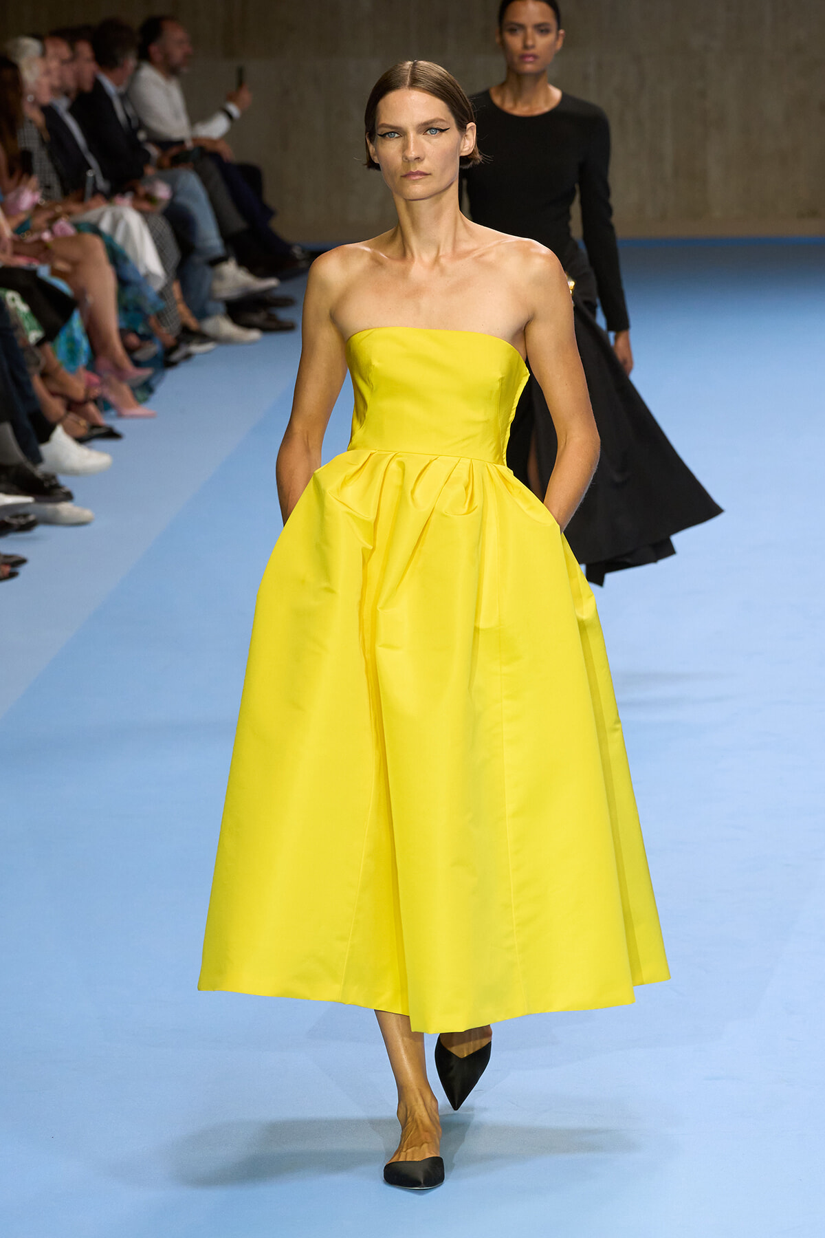 Model walks a yellow strapless ball gown with a fitted bodice and full midi-length skirt on a blue runway, audience on sides