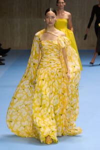 Model walking a runway in a yellow floral gown with a flowing cape, off-shoulder bodice, and matching yellow heels.